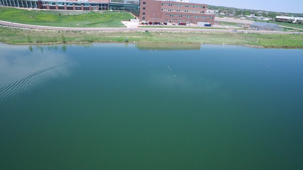 Aerial view over green-blue lake with brick buildings and a road beyond the shore.