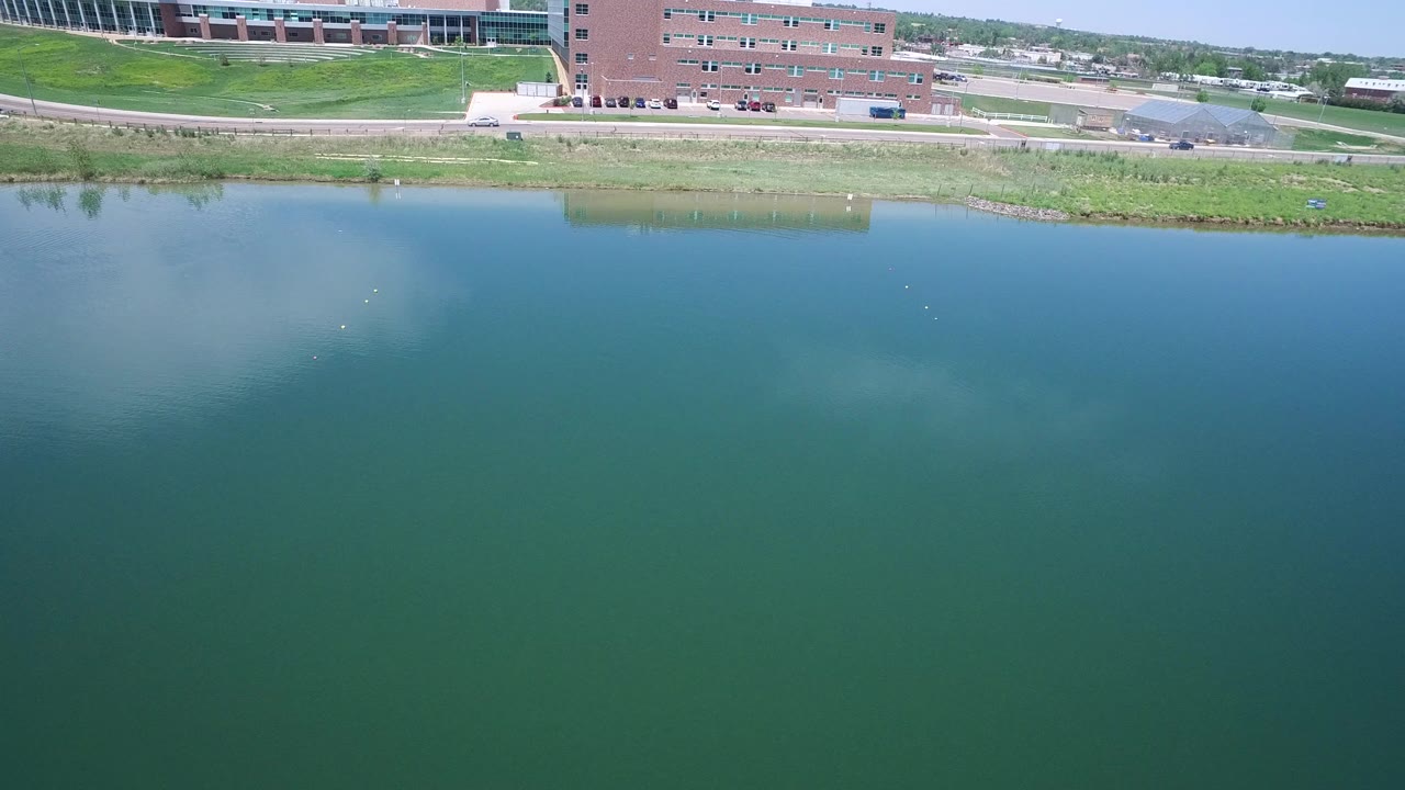 Opening aerial frame in Westminster, Colorado, with blue-green water dominating the view and brick buildings, grass, road, and cars along the far shoreline.