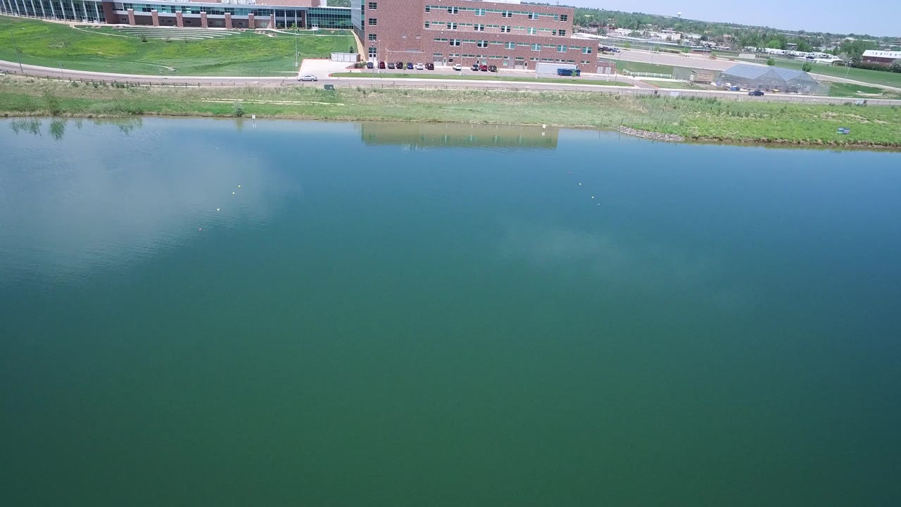 Second aerial frame in Westminster, Colorado, showing a broad reflective water surface below a grassy bank, a road with light traffic, and modern buildings at the top of frame.