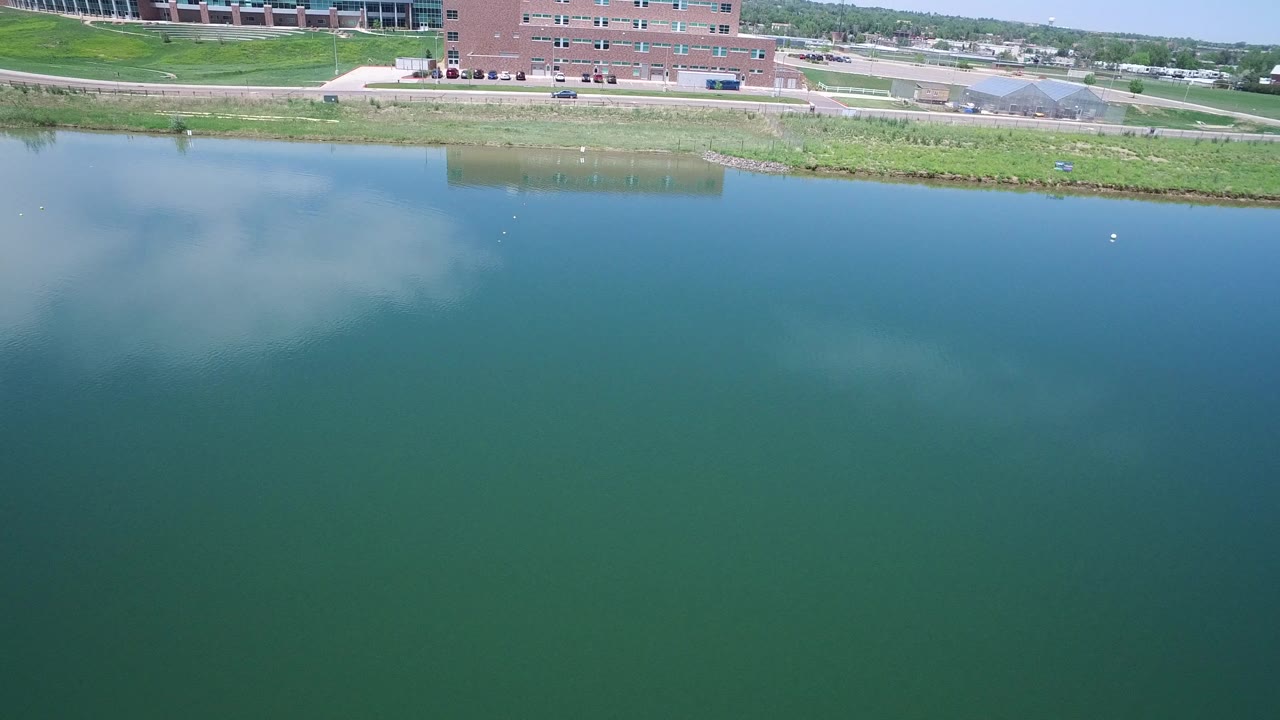 Opening view of blue green water in Westminster, Colorado with a brick waterfront building, roadway, parked cars, grassy bank, distant trees, and no visible people.