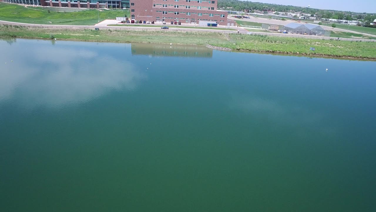 A second moment shows reflective blue water in Westminster, Colorado below a grassy shore, brick building, roadway traffic, parked cars, and no visible people.