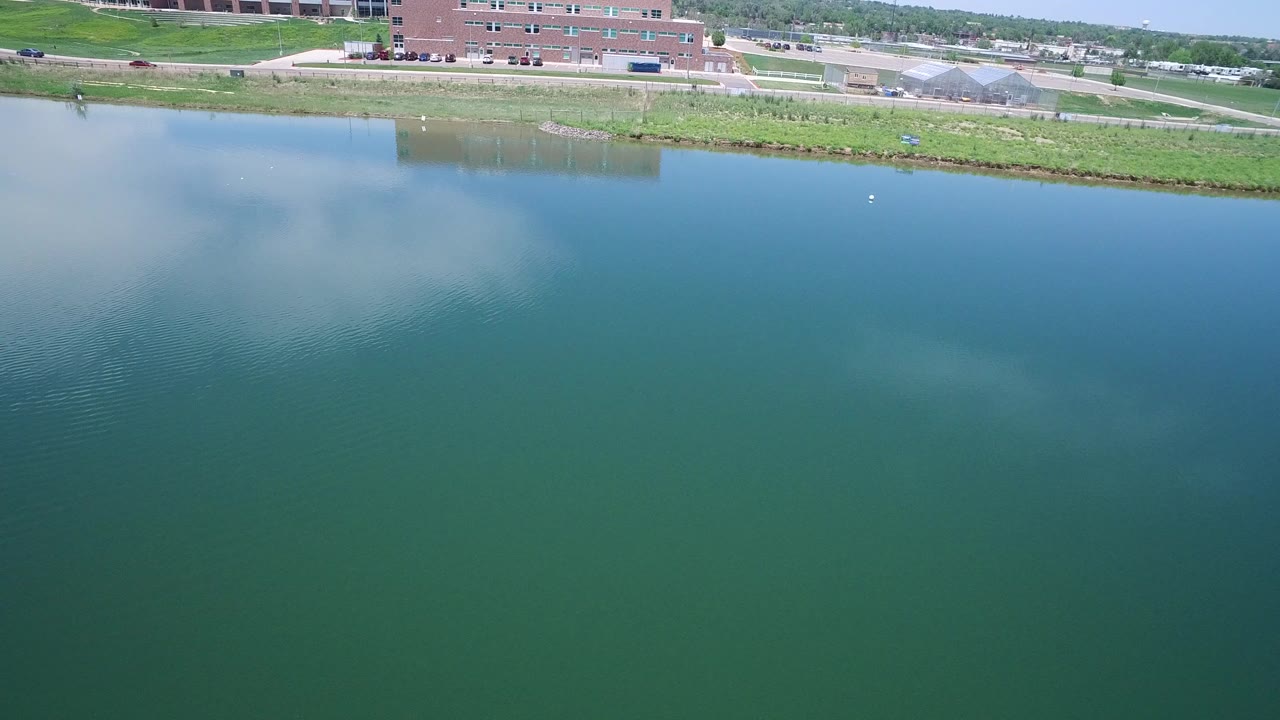 Aerial view of calm lake water beside a grassy shoreline and brick buildings.