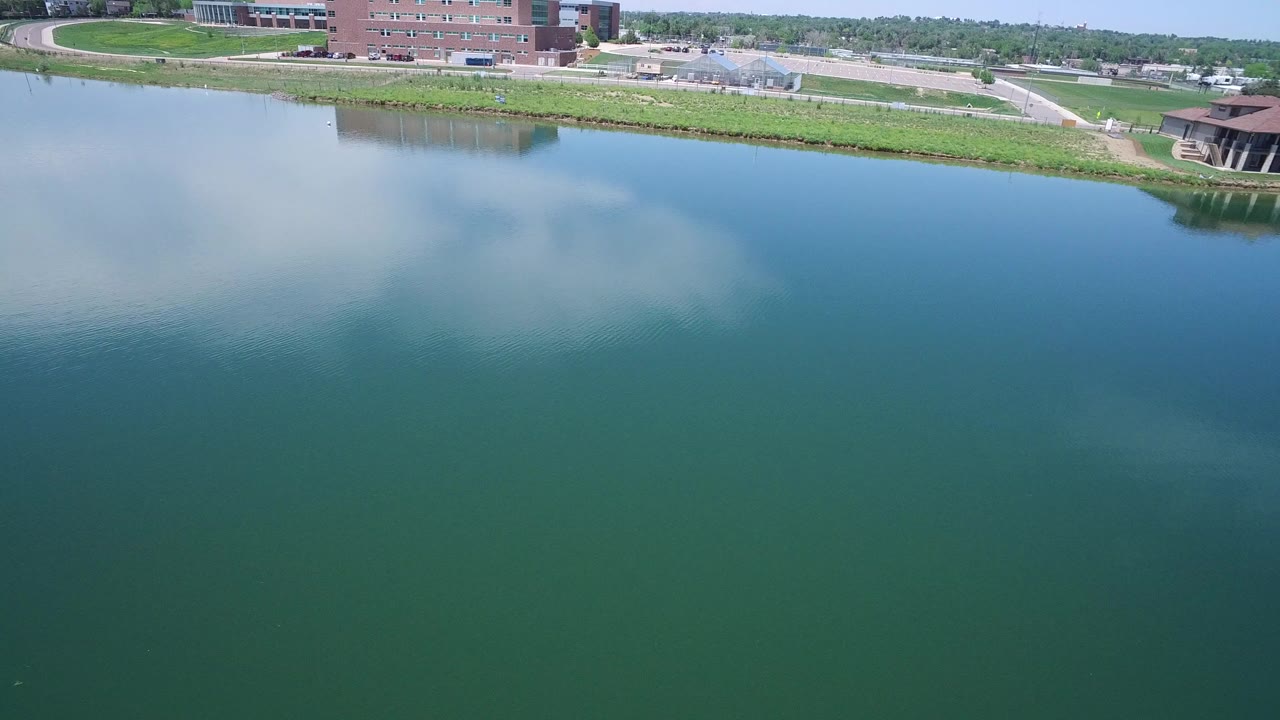 Aerial view in Westminster, Colorado looking over blue-green water toward a grassy bank, a brick building, paved paths, and a small pavilion structure, with no visible people.