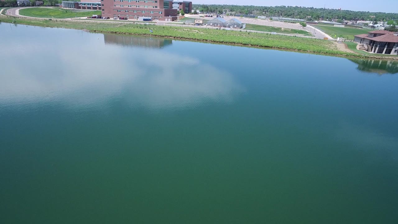 Blue water with cloud reflection below a grassy bank and distant buildings