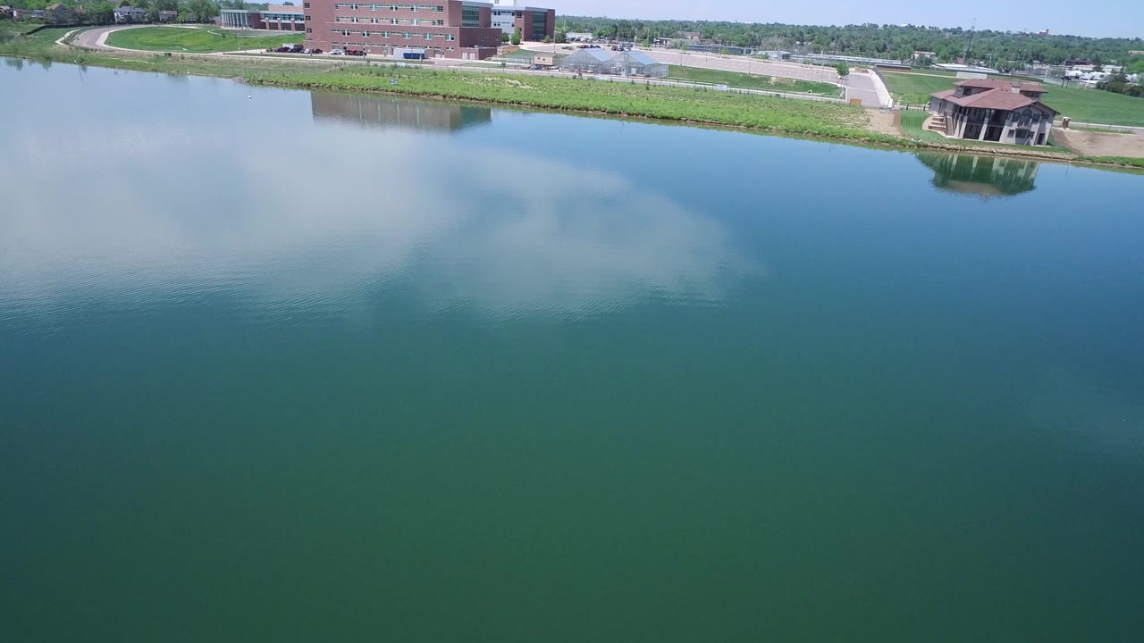 Aerial view over the Westminster lake with reflected clouds centered on the water, a pavilion near the right shoreline, brick buildings behind it, and no visible people.