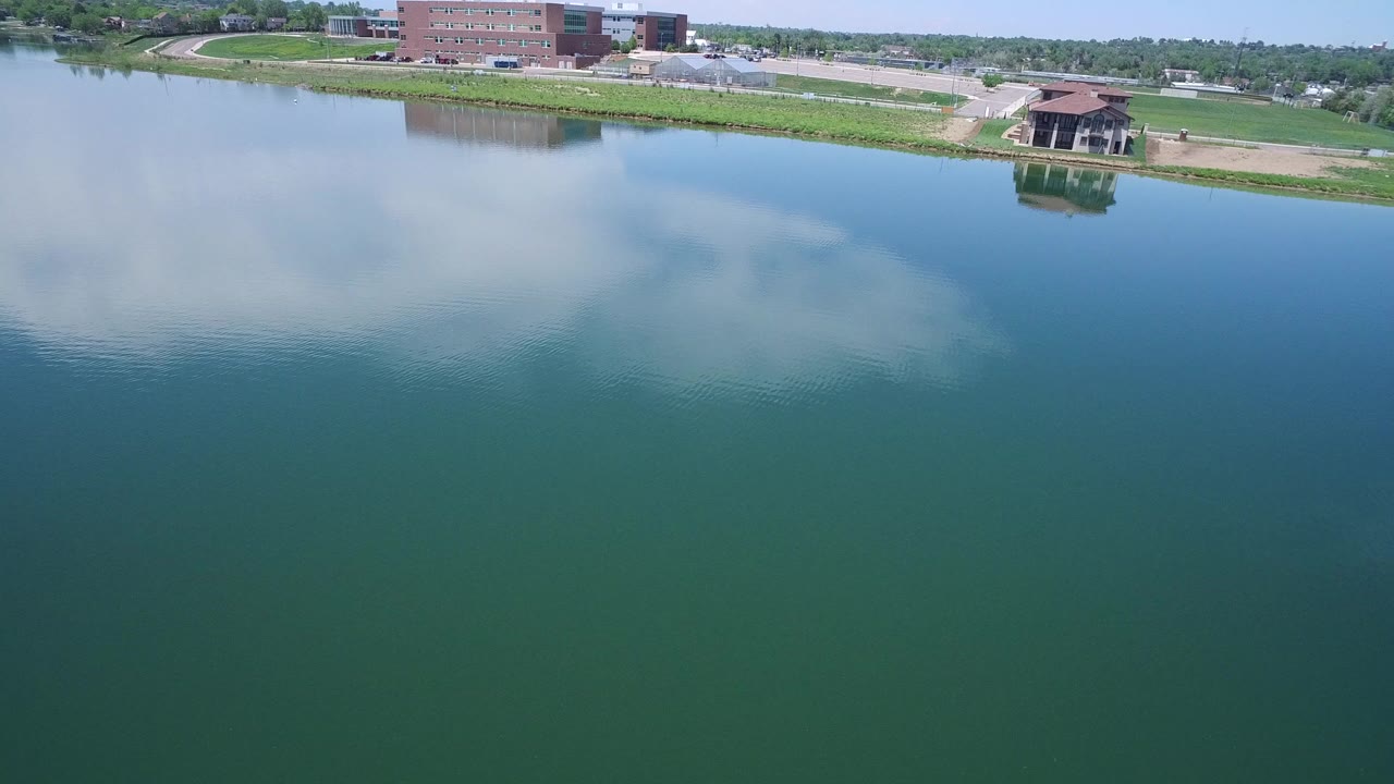 Aerial view over a Westminster, Colorado lake with blue-green water, cloud reflections, a grassy shoreline, brick buildings, a pavilion, and no visible people.