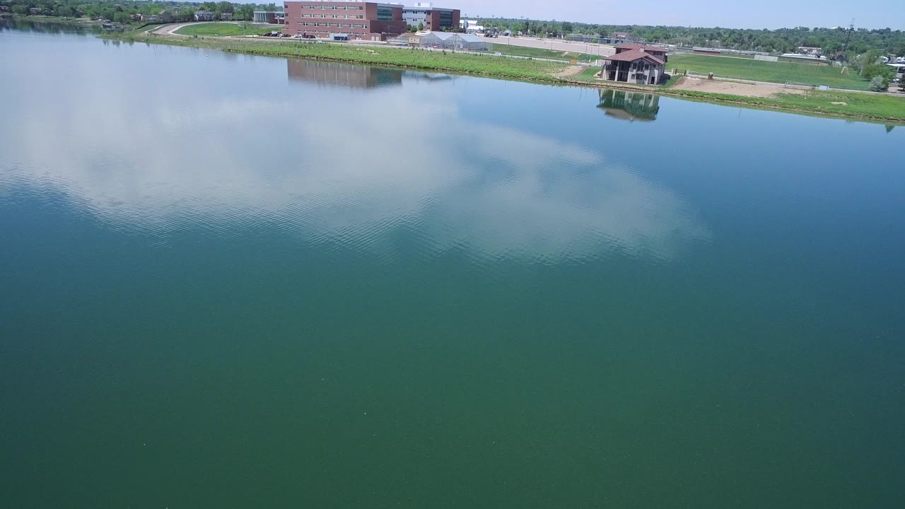 Opening frame of a Westminster, Colorado lake view, with broad blue-green water, cloud reflections, grassy shoreline, distant buildings, and no visible people.
