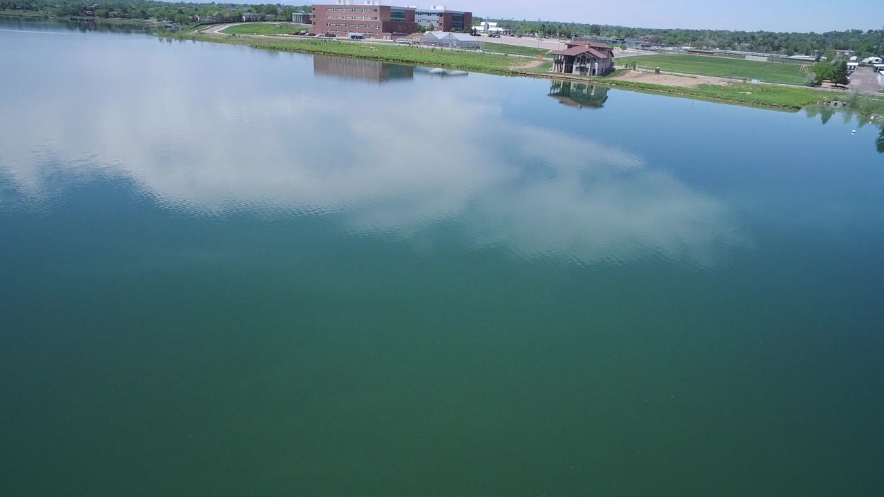 Aerial view over a blue-green lake with cloud reflections and a grassy shoreline.