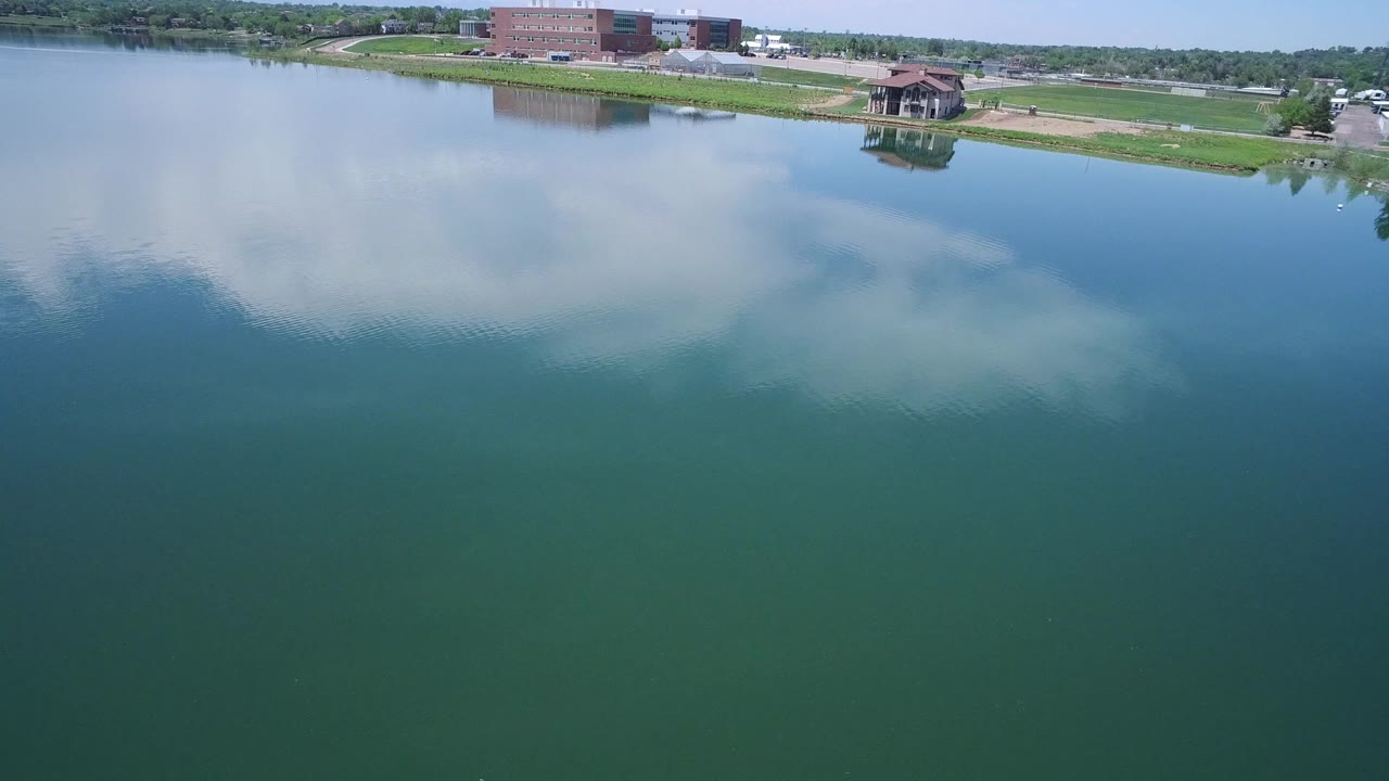 Aerial frame in Westminster, Colorado showing the lake surface dominating the view, with white clouds reflected in the water and the pavilion and brick building near the upper shoreline, with no visible people.