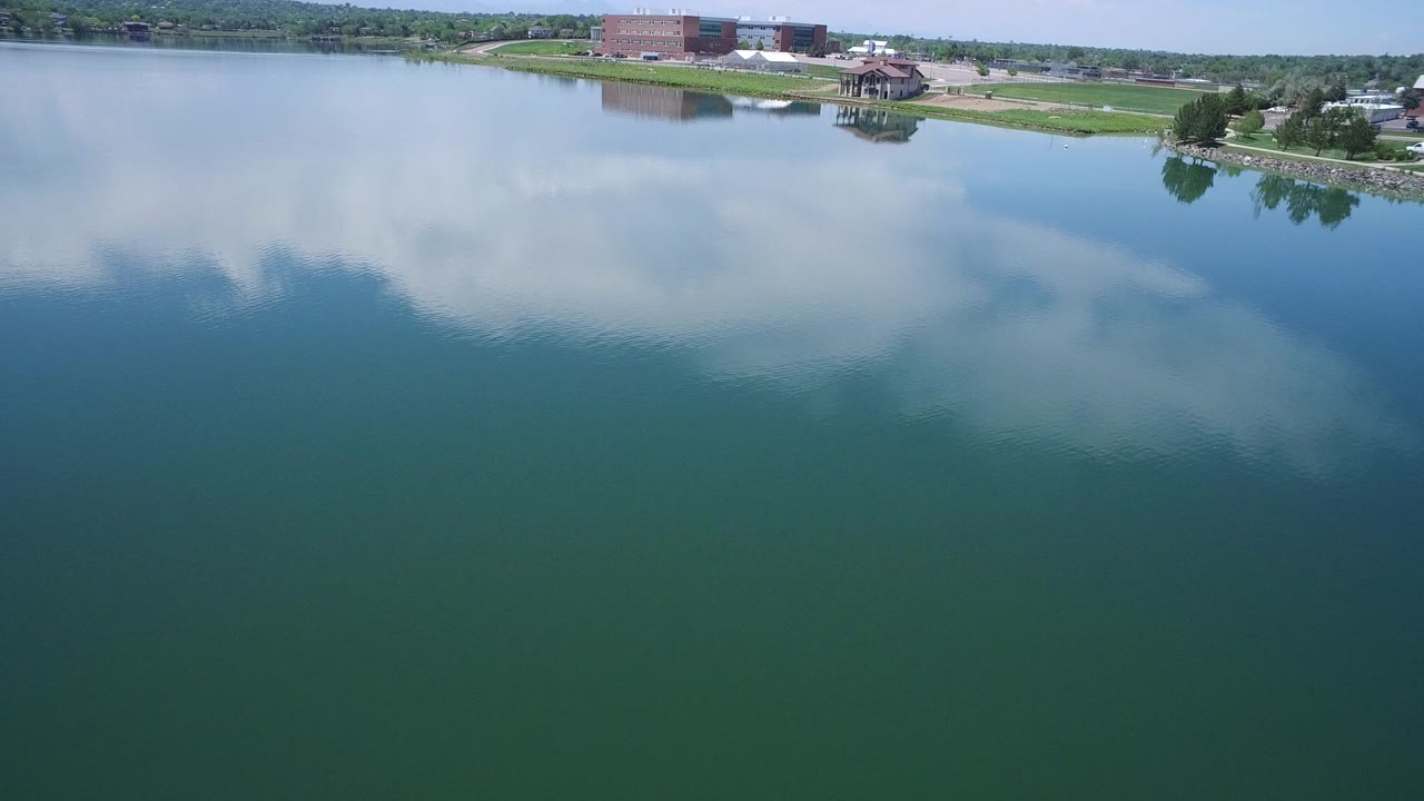 A high view over blue-green lake water in Westminster, Colorado shows broad cloud reflections, a grassy bank, trees, a small pavilion, distant buildings, and no visible people.