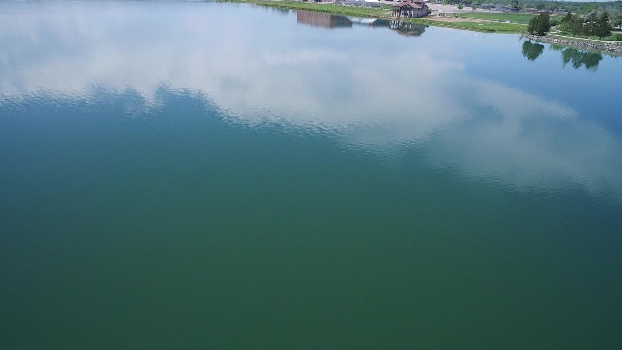 Green lake water with cloud reflections and a narrow shoreline with houses at the top.