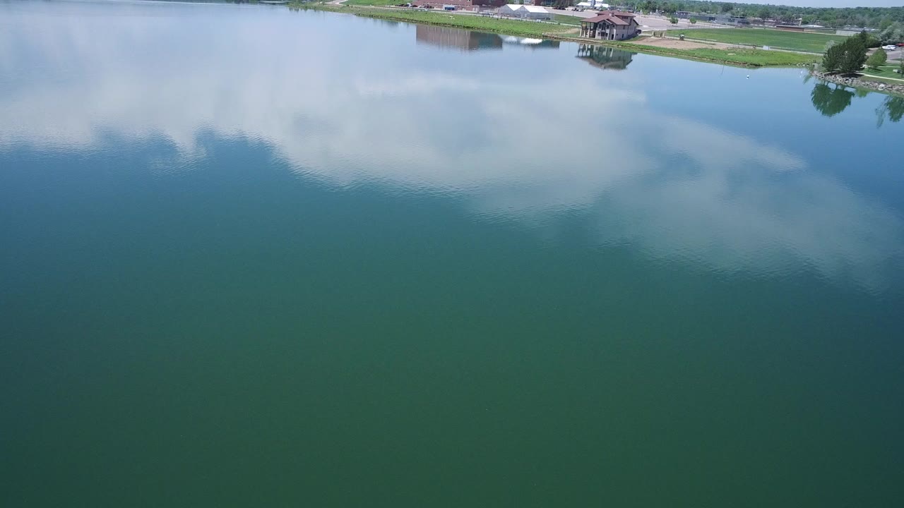 Aerial frame over a large reflective body of water in Westminster, Colorado, with cloud reflections across the surface, a grassy shoreline at the top, distant buildings, and no visible people.