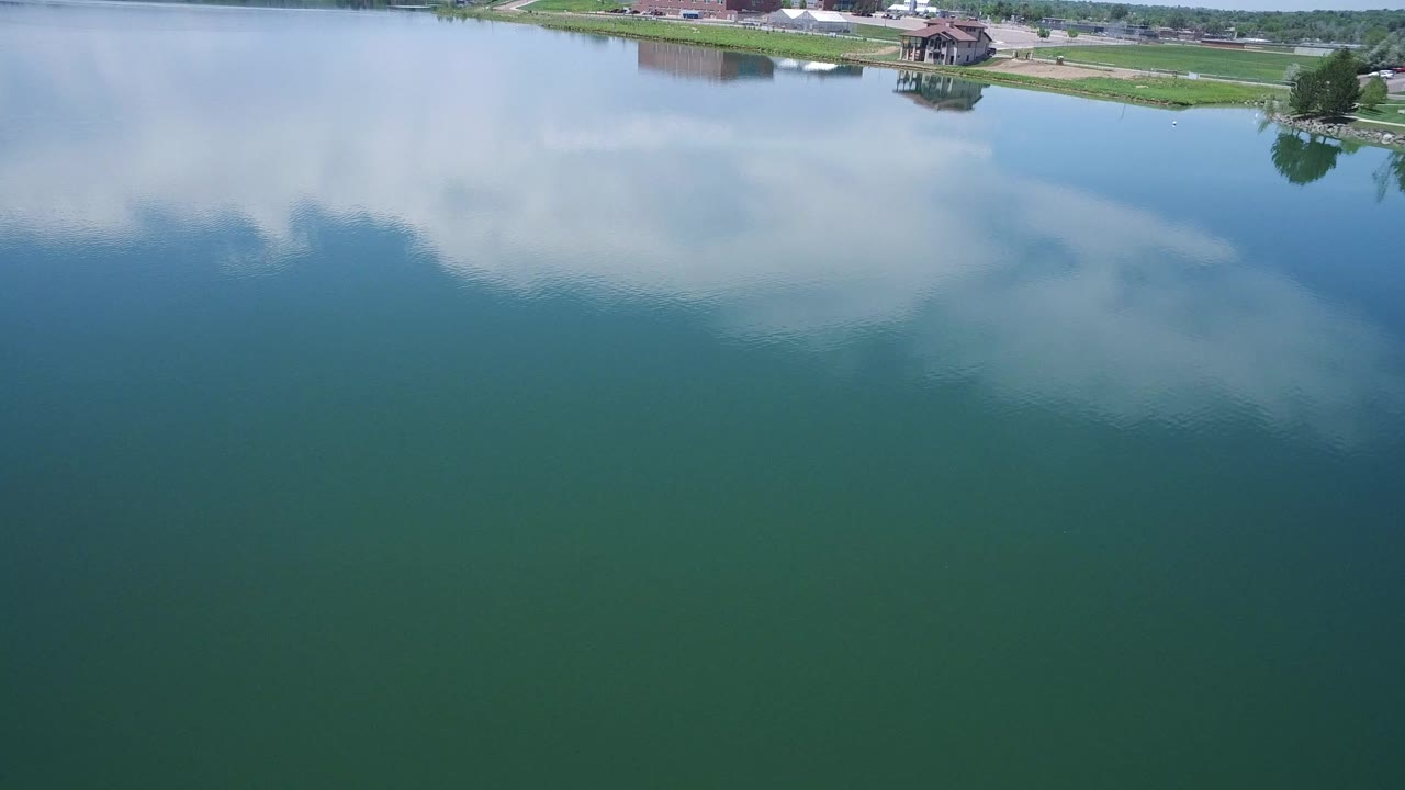 Aerial water surface with cloud reflection and a distant shore with grass and structures.