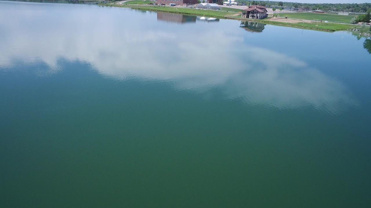Aerial view of green lake water with cloud reflections and shoreline at the top.