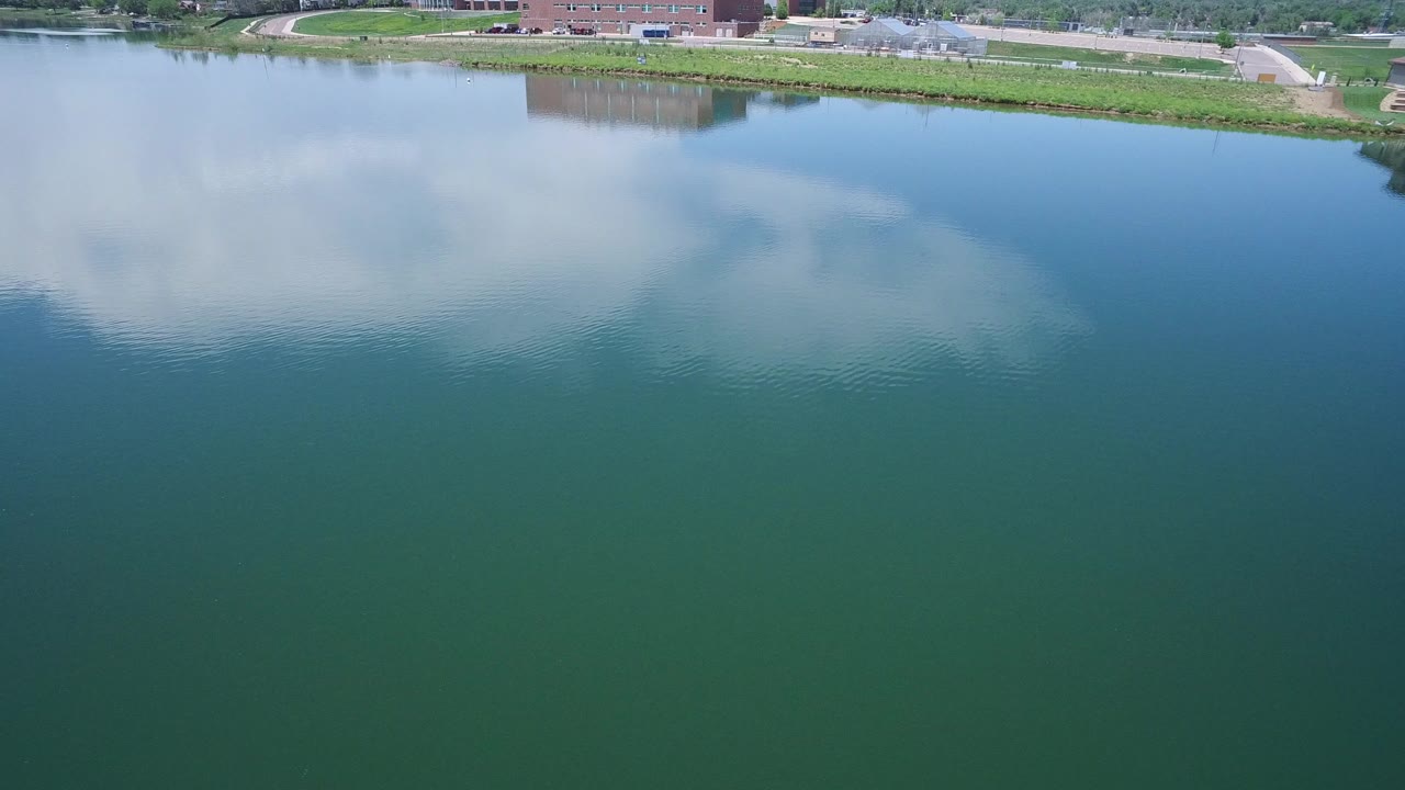 Opening frame of a Westminster, Colorado lake view with cloud reflections across blue-green water, a grassy shore, roads, and low buildings in the distance, with no visible people.