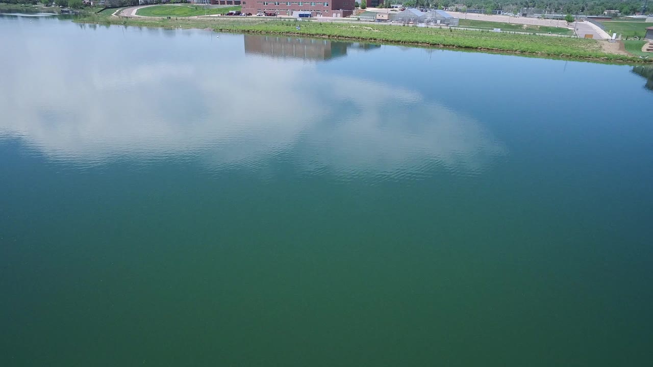 Second frame shows the reflected cloud mass shifted across the Westminster lake surface, with the grassy bank, roadway, and distant buildings still near the upper edge, with no visible people.