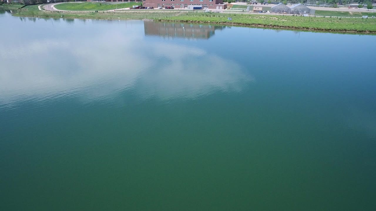 Aerial view in Westminster, Colorado showing blue-green water with a broad cloud reflection, faint ripples on the left, a grassy bank, distant paths, and no visible people.