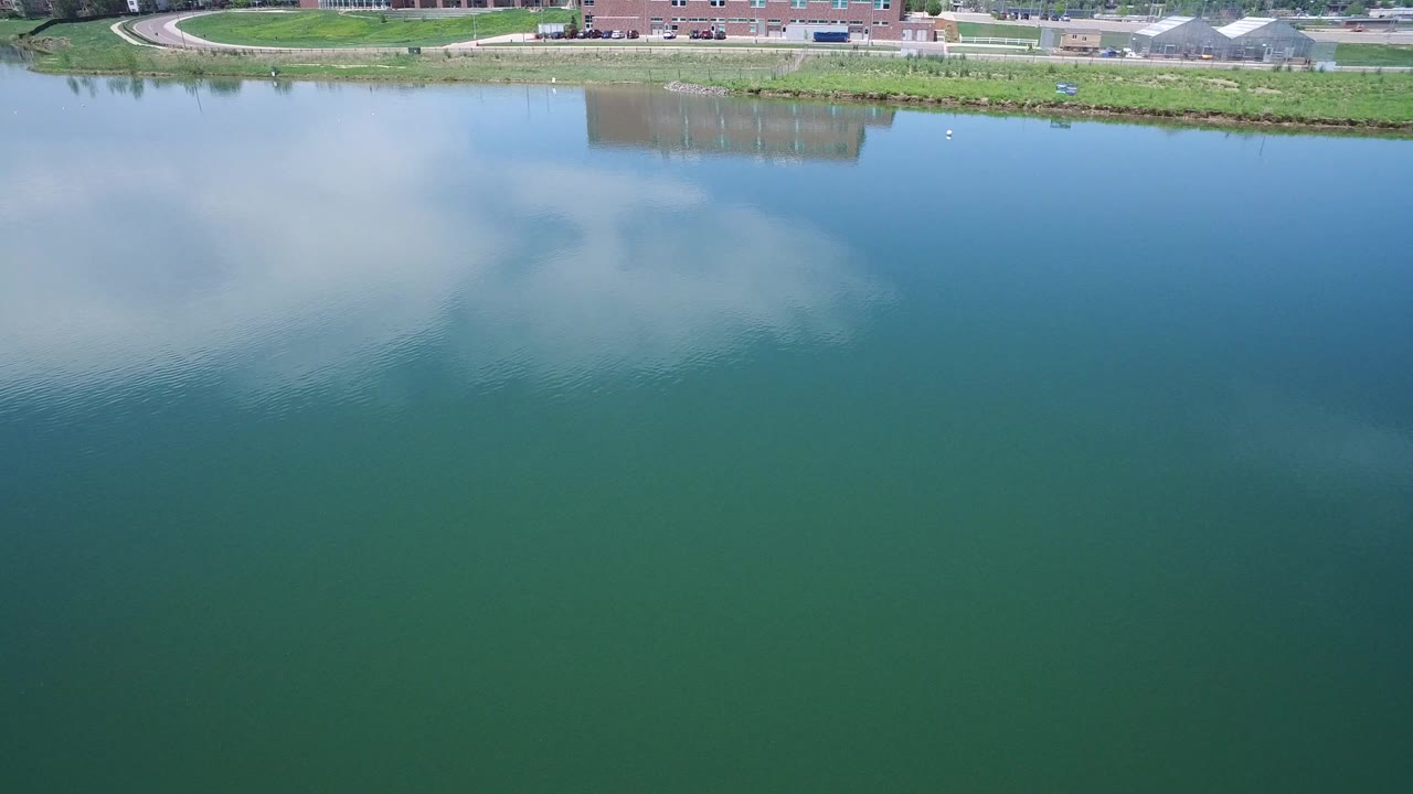 Aerial view of green-blue water with cloud reflections and a shoreline building at the top edge.