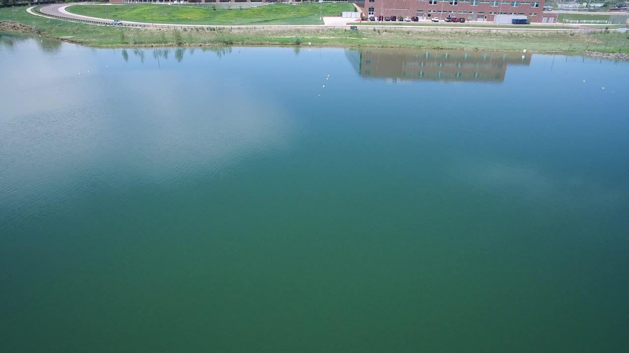 Aerial view over blue-green pond water with a grassy shoreline at the top edge.
