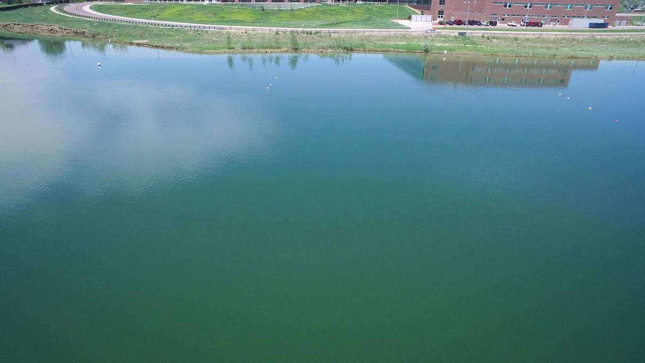 Aerial frame in Westminster, Colorado showing green-blue water, faint cloud reflections, a grassy shoreline, a curving path, a brick building, parked cars, and no visible people.
