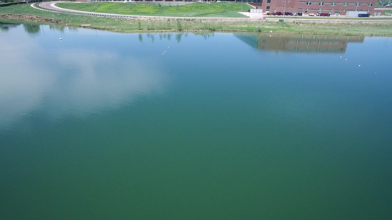 Aerial Westminster, Colorado view with broad teal water in the foreground, soft cloud reflection on the left, a thin grassy bank, path, brick building, parked cars, and no visible people.