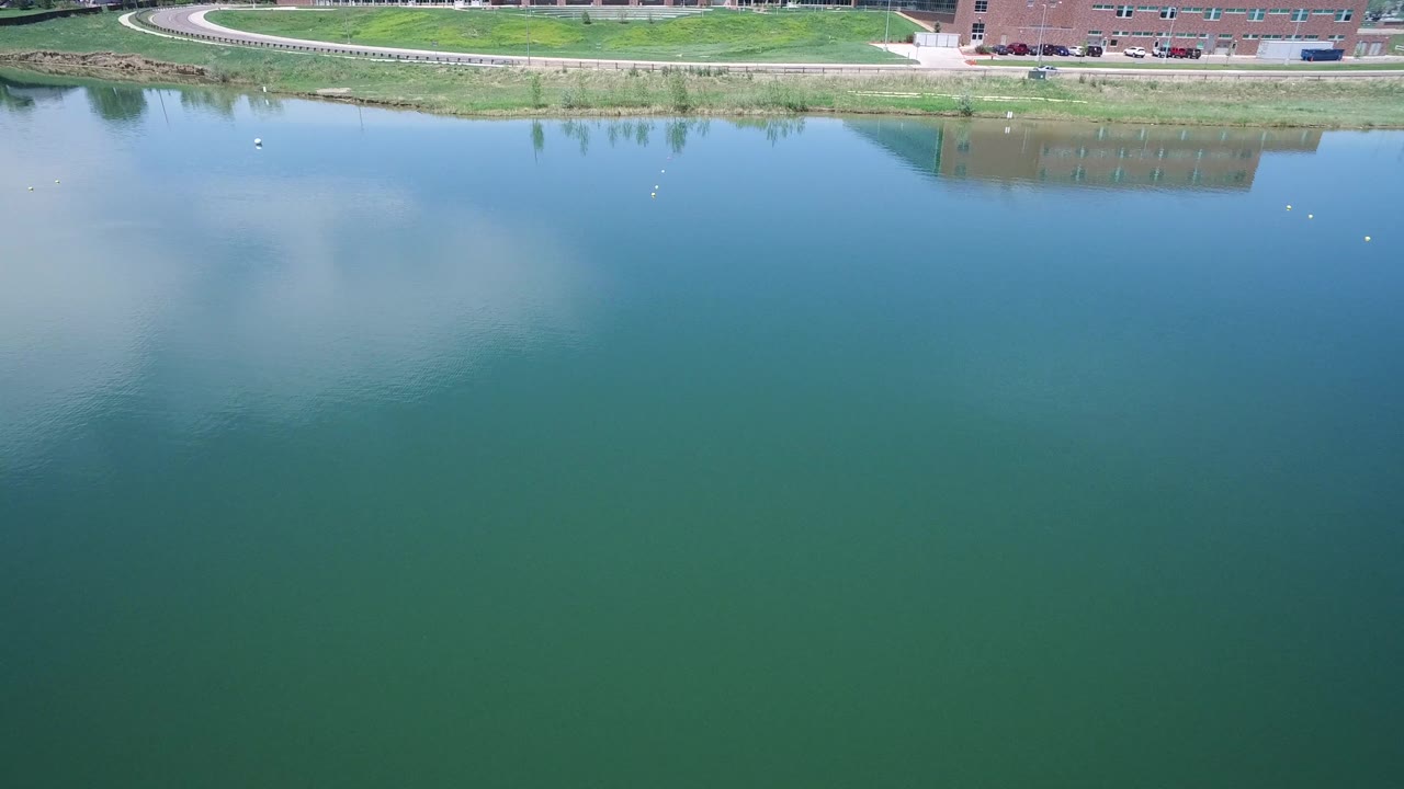Wide aerial view across teal water toward a grassy bank and parked cars.