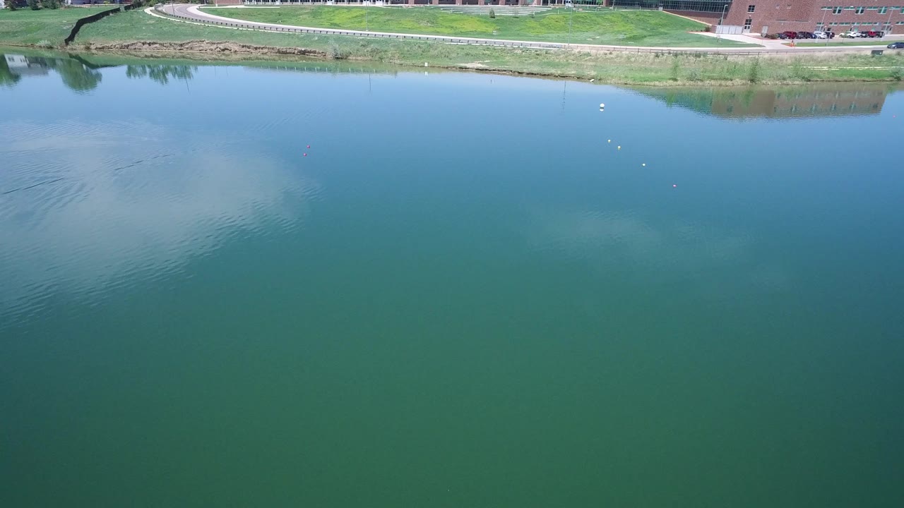 Westminster, Colorado view over calm blue-green water with a grassy bank, small vehicles along the road, a brick building at the edge, and no visible people.