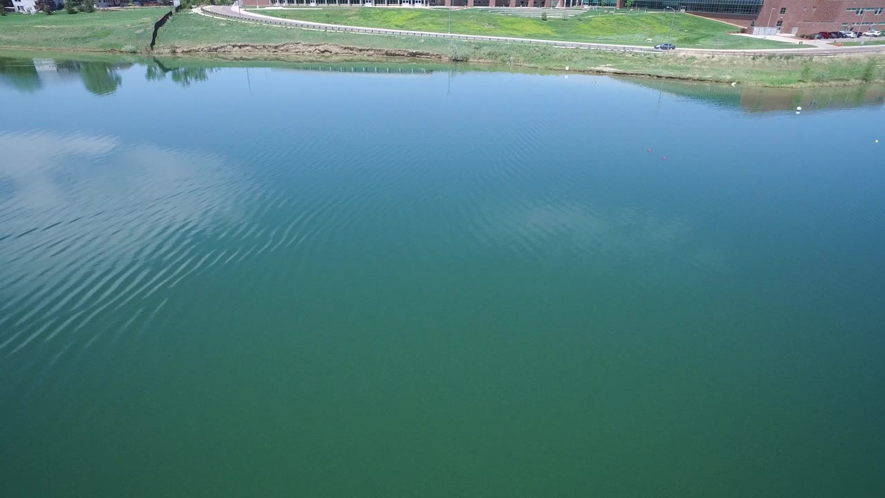 Wide elevated view of a blue-green Westminster, Colorado lake with ripples crossing the water, a grassy shoreline and road at the top, and no visible people.