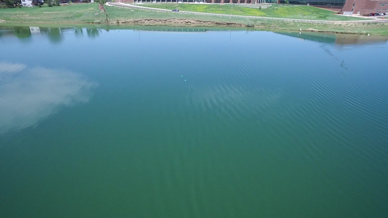 Aerial view over green-blue water with a grassy bank and buildings at the top edge.