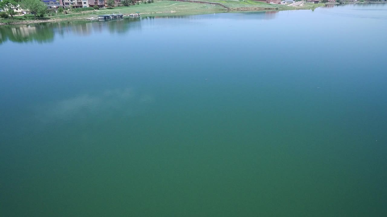 Aerial view over blue-green lake water with a distant grassy shoreline.