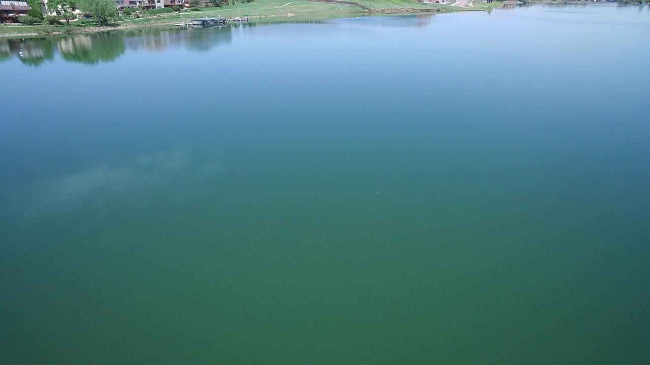 Opening frame of blue green lake water in Westminster, Colorado, with a thin grassy shoreline, trees, docks, houses, and no visible people.