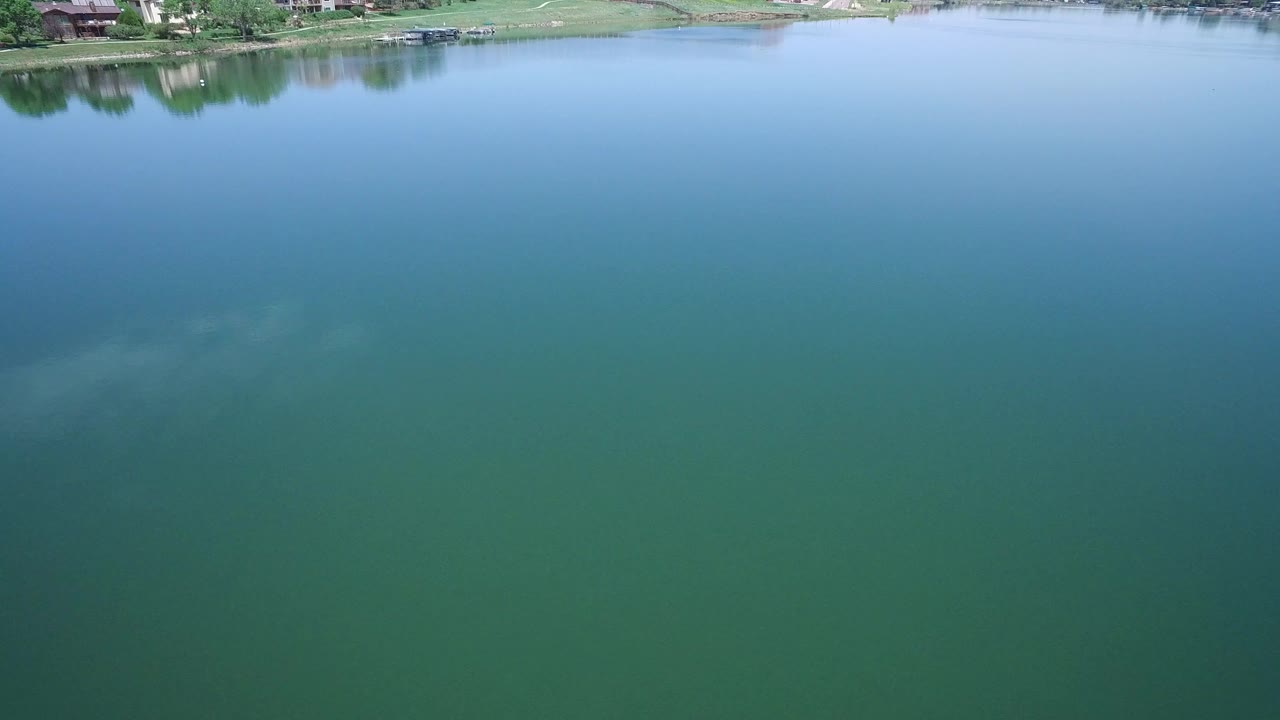 Second view across the Westminster, Colorado lake, with smooth blue water in the center, green water in the foreground, distant homes near the shore, and no visible people.