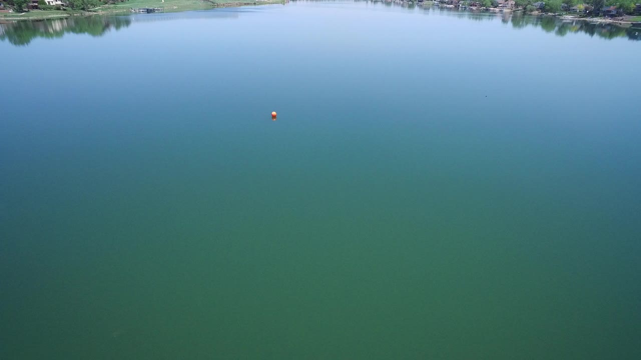 Aerial view over a calm green-blue lake with a small red marker and distant shoreline.