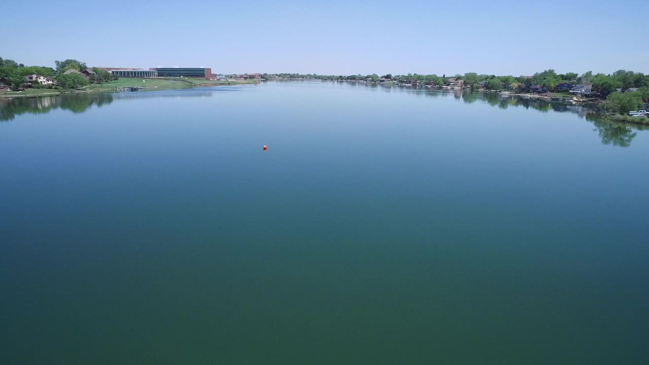 Calm water dominates the frame in Westminster, Colorado, with a tree-lined residential shoreline across the distance, a large building left of center, and no visible people.