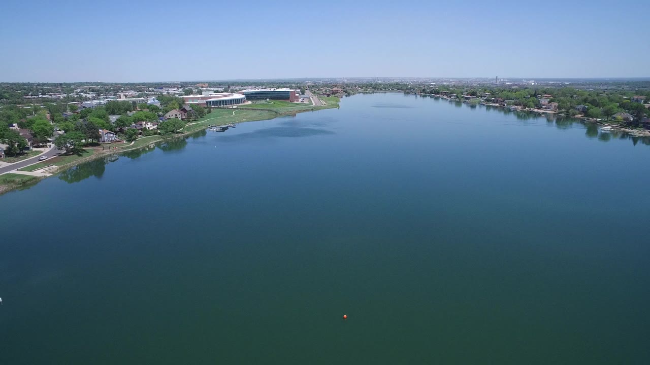 Aerial view in Westminster, Colorado over blue-green water with shoreline homes, trees, roads, and a large white-roofed building in the distance, with no visible people.