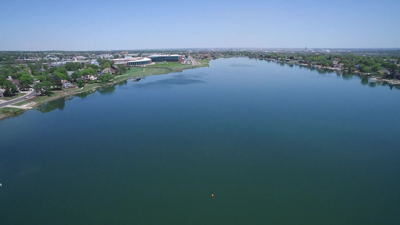High aerial frame in Westminster, Colorado showing broad blue-green water, a tree-lined residential shore, distant buildings, and no visible people.