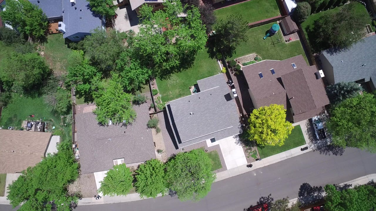 Straight-down aerial view of Westminster, Colorado homes with gray and brown roofs, green yards, sidewalks, parked cars, and no visible people at the start of the clip.