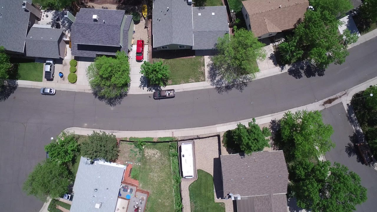 Overhead aerial frame of Westminster, Colorado rooftops, front yards, sidewalks, multiple parked vehicles, and tree shadows across a residential street, with no visible people.