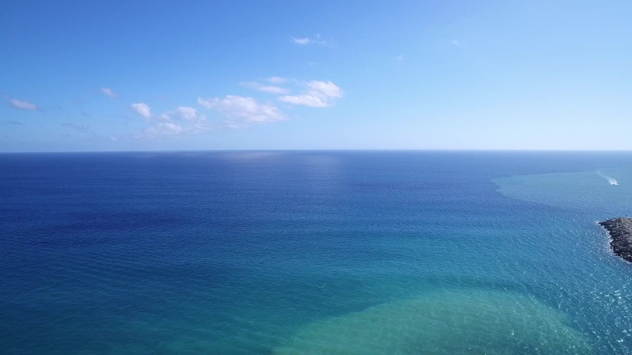Opening frame over Playita, Baja California Sur water, with deep blue ocean, turquoise shallows near the bottom, a clear horizon, small clouds, and no visible people.
