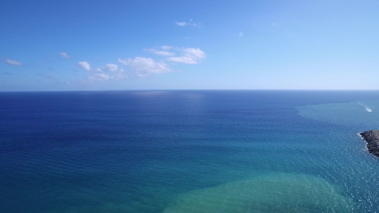 Aerial view over Playita, Baja California Sur, Mexico, showing deep blue ocean, turquoise shallows, scattered clouds, and no visible people at the start of the clip.