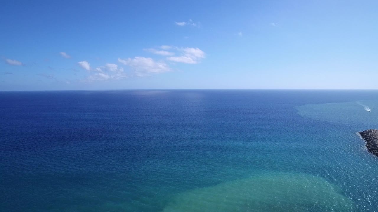 Wide aerial frame over Playita with the dark blue sea spanning the background, pale turquoise water below, a small rocky edge at right, and no visible people.