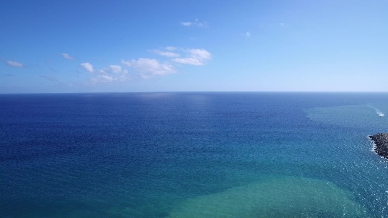 Opening frame over Playita, Baja California Sur, Mexico, showing blue ocean, lighter turquoise shallows in the lower frame, a small cloud cluster, and no visible people.