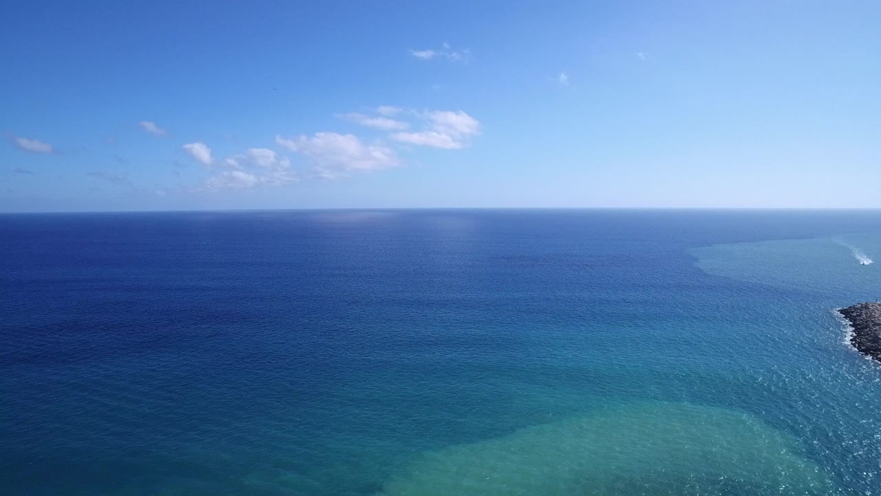 Playita coastal water seen from above, with deep blue sea, turquoise bands near the bottom, a dark rocky shoreline edge at right, and no visible people.