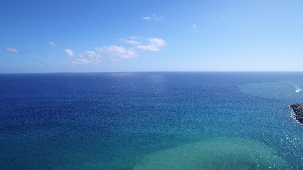 Aerial frame over Playita water showing deep blue ocean, pale turquoise shallow water, a small white surf break near a dark rocky edge, blue sky, and no visible people.
