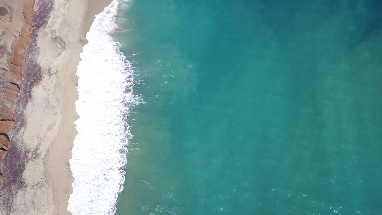 High overhead view of Playita, Baja California Sur with sand along the left edge, a bright white surf band, turquoise water to the right, and no visible people.
