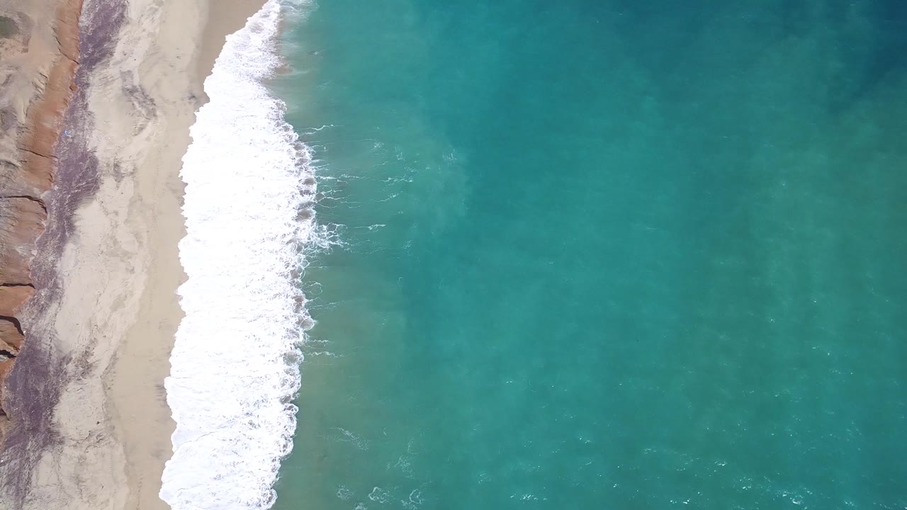 Overhead Playita shoreline frame with the foamy wave line running down the left side and a broad field of teal ocean water filling the right side, no visible people.