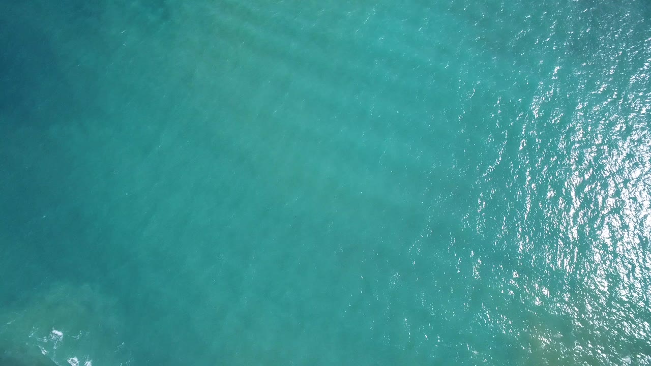 Overhead view of turquoise seawater in Playita, Baja California Sur, with rippled surface, bright reflection to the right, and no visible people.