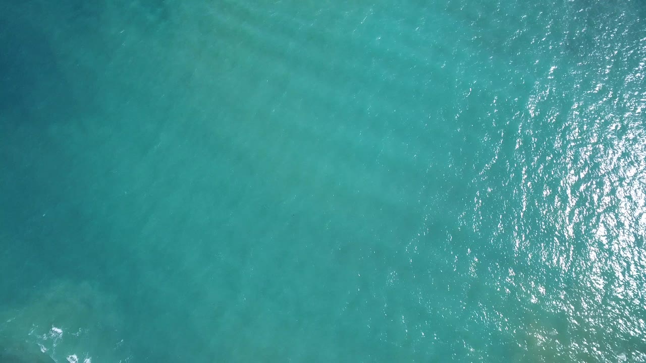 Aerial-looking frame over Playita blue sea, showing darker underwater tones, soft ripples, a small white foam mark near the lower left, and no visible people.