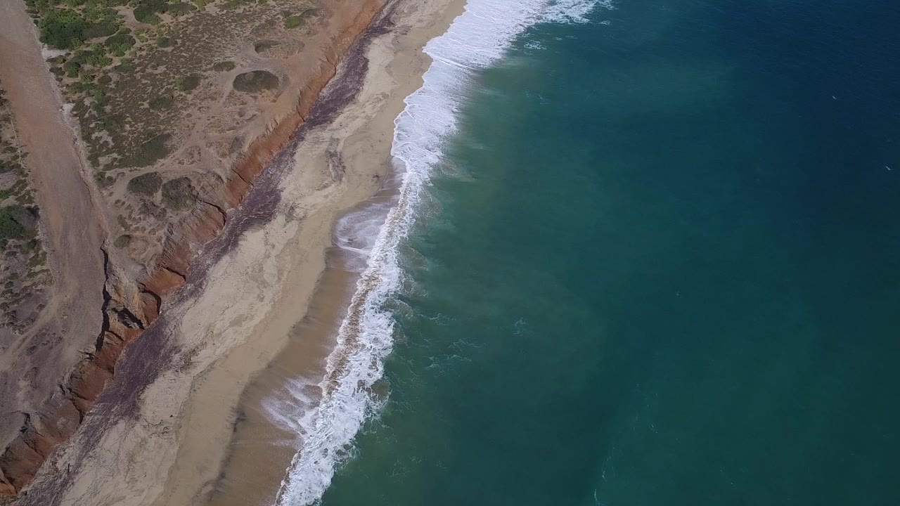 Aerial frame over Playita with the surf line shifted down the beach, blue-green water on the right, scrubland on the left, and no visible people.