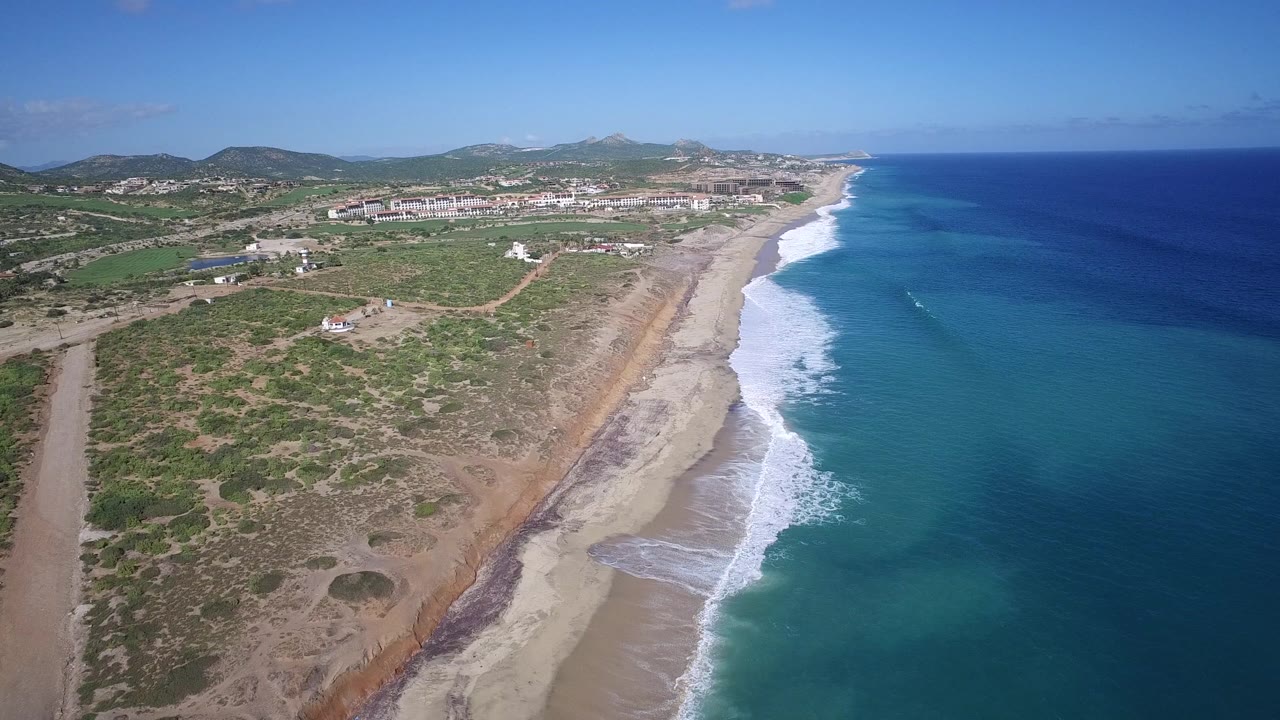 Drone-style view of Playita beach with white surf curling along the shore, turquoise shallows, dry scrubland and tracks inland, distant resort buildings, hills, and no visible people.